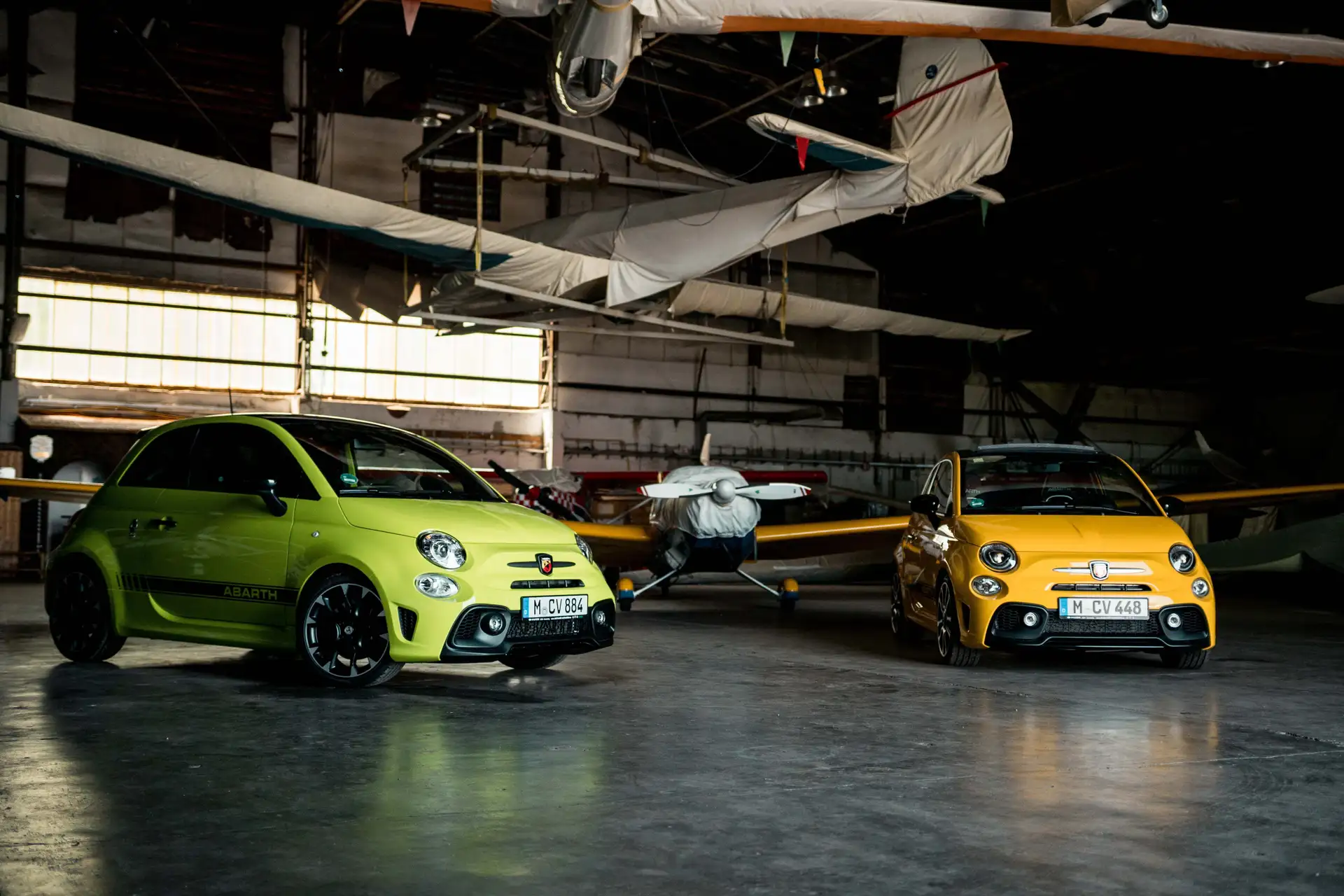 A green Abarth 595 Competizione stands next to an Abarth 595 Turismo Cabrio in a hangar.